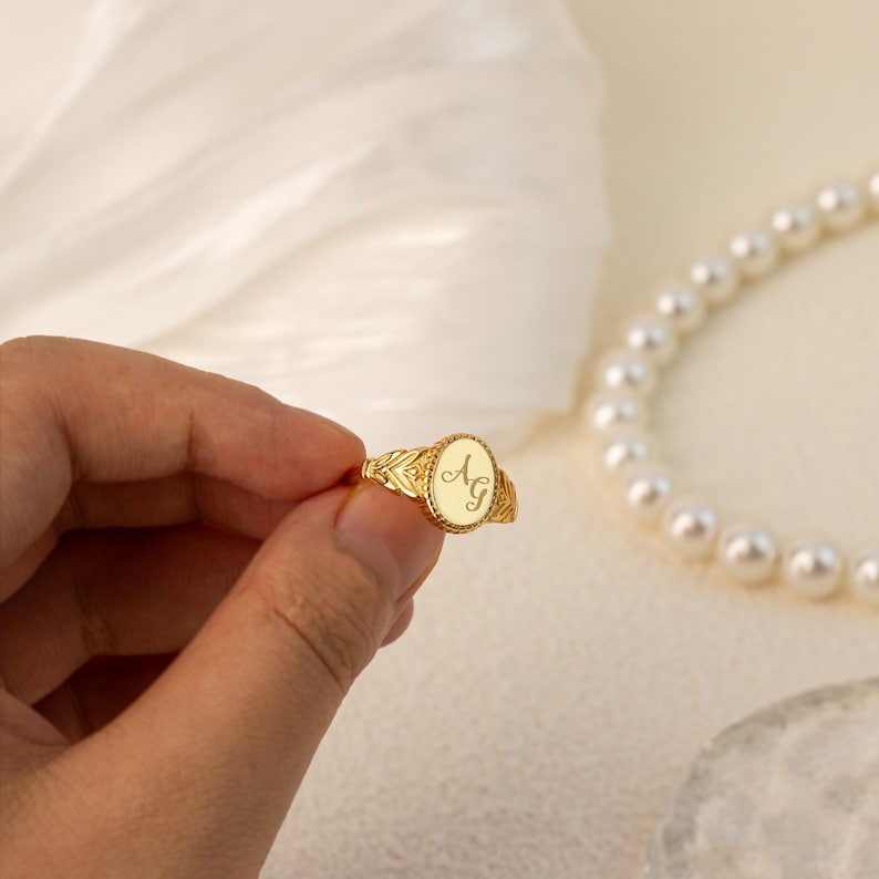 May include: A gold signet ring with an oval-shaped top featuring the initials "AG" engraved in a cursive script.