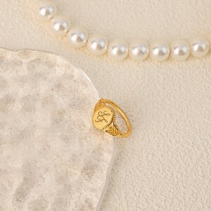 May include: Gold signet ring with a round, engraved top featuring the initials "LF". The ring has a delicate, ornate design.