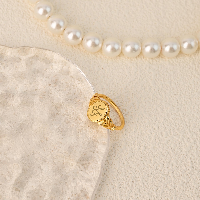 May include: Gold signet ring with a round, engraved top featuring the initials "LF". The ring has a delicate, ornate design.