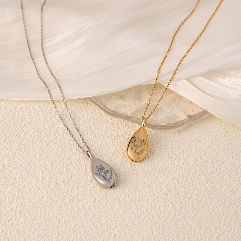 May include: Two teardrop-shaped pendant necklaces. One is silver-toned, the other gold-toned. Each pendant features a floral design and a name engraved on the front. The necklaces are displayed on a light-colored surface.