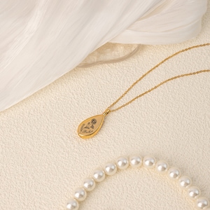 May include: A gold-colored necklace with a teardrop pendant featuring a floral design and the word "Beth". The necklace is on a delicate chain, and a strand of pearls is visible in the foreground.
