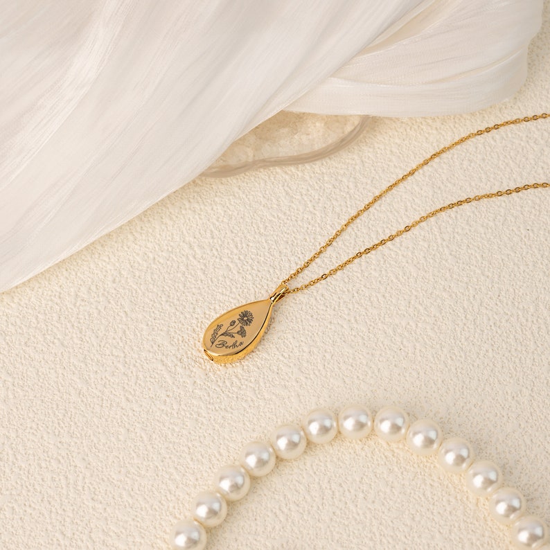 May include: A gold-colored necklace with a teardrop pendant featuring a floral design and the word "Beth". The necklace is on a delicate chain, and a strand of pearls is visible in the foreground.