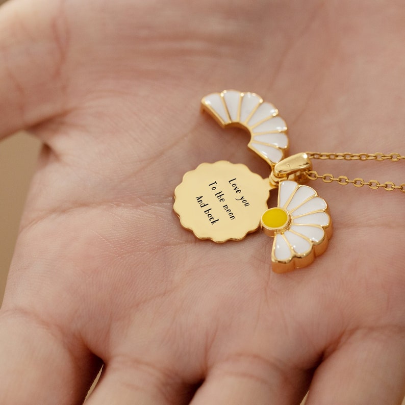Hand holding an engraved white flower necklace that opens up and reads Love you to the moon and back
