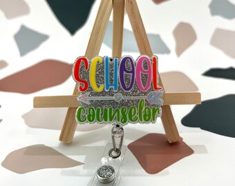 May include: A silver glitter badge holder with a rainbow colored "SCHOOL COUNSELOR" text and a silver arrow with a heart. The badge holder is on a small wooden easel.