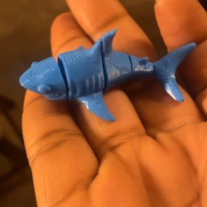 May include: A blue plastic toy shark with segmented body parts. The shark is being held in a hand.