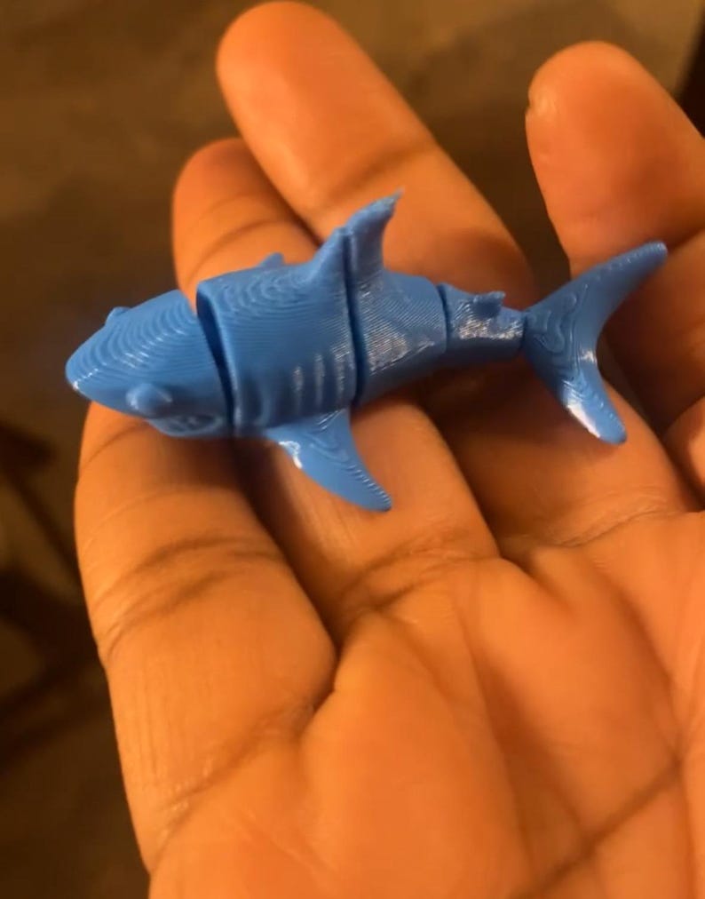 May include: A blue plastic toy shark with segmented body parts. The shark is being held in a hand.