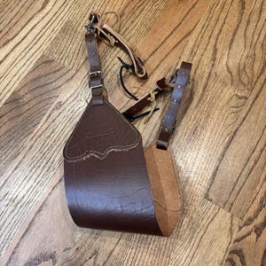 Può includere: Un faretra da fianco da tiro con l'arco in pelle marrone con un bordo superiore decorativo e cinghie regolabili. La faretra è progettata per contenere le frecce e ha un interno beige a contrasto. Le cinghie hanno fibbie metalliche e una clip.