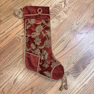 May include: Burgundy Christmas stocking with gold floral embroidery. The stocking has a velvet texture, a gold rope trim, and a small pocket. A gold tassel and bells hang from the bottom.