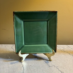 May include: A square, emerald green ceramic plate with a slightly textured surface. The plate has a dark brown rim and is displayed on a decorative, gold-colored stand. The plate is a solid color and has a glossy finish.