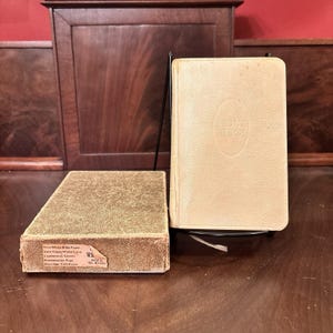 May include: A white Holy Bible with a textured cover stands upright on a black metal display stand next to its matching gold-toned box. The box features a label listing Thin White Bible Paper, Gold Edges, White Lims, Leatherette Covers, Presentation Page, and Marriage Certificate.