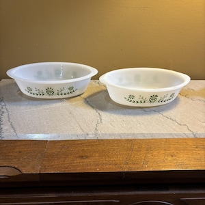 May include: Two white oval-shaped Pyrex baking dishes with green floral designs. The dishes have handles on each side and are sitting on a light-colored surface. The floral pattern includes stylized flowers and leaves.