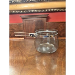 May include: A clear glass saucepan with a transparent handle and a silver-colored metal rim. The saucepan is sitting on a wooden surface.