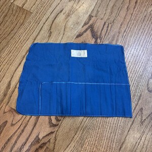 May include: A rectangular blue fabric tool roll featuring a series of vertical stitched pockets for storage. The item includes a small white rectangular label centered near the top edge and is constructed with reinforced blue stitching along the borders.