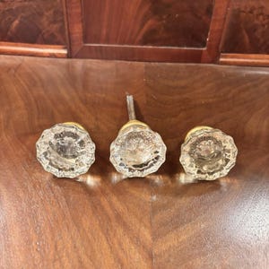 以下が含まれることがあります： ファセットデザインと真鍮金具を備えた、透明なガラス製のキャビネットノブ3つ。木製の表面に配置されており、ヴィンテージ感を強調しています。家具の修復や装飾に適しています。