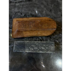 May include: A brown leather case with the embossed text "Smith's" and a rectangular sharpening stone. The case is stitched along the edges. The stone is dark grey and rectangular.