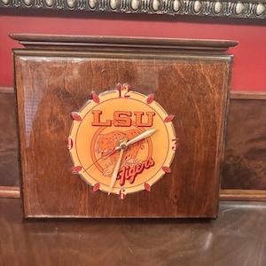 May include: A wooden wall clock featuring the LSU Tigers logo. The clock face is orange with the team's tiger mascot and the words "LSU Tigers." The clock has a dark brown wooden frame and gold-colored clock hands.