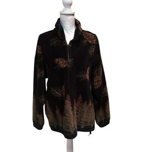 Puede incluir: Chaqueta negra de forro polar con cremallera y cuello alto. La chaqueta presenta un estampado de bosque y piñas en tonos marrones y verdes. La chaqueta tiene un cordón en la parte inferior y se muestra en un maniquí.