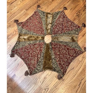 May include: A decorative tree skirt with a starburst design. The skirt features a rich burgundy and olive green floral pattern, accented with brown faux fur and tassels. The centre has a circular opening for the tree trunk.