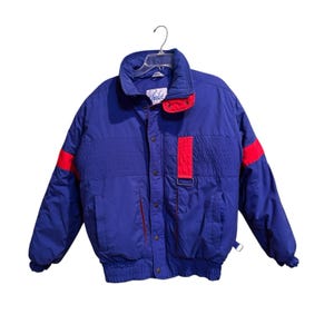 Puede incluir: Chaqueta de invierno azul y roja con cuello alto y diseño acolchado. La chaqueta tiene detalles rojos en los hombros y un panel rojo en la parte delantera. La chaqueta está colgada de una percha transparente.