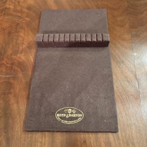 Puede incluir: Un paño marrón Reed & Barton anti-deslustre para cubiertos de plata. El paño rectangular tiene una sección elevada con ranuras. El logotipo de Reed & Barton es visible.