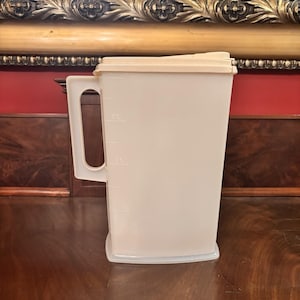 Puede incluir: Una jarra de plástico beige con asa y tapa. La jarra tiene marcas de medición en el lateral. El asa es rectangular y está unida al lateral de la jarra. La tapa es de un tono beige más claro.