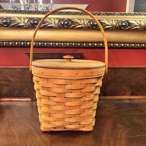 Peut inclure: Un panier tissé brun clair avec un couvercle et une poignée en bois. Le panier est de forme ronde et est fait de lanières de bois tissées. Le couvercle est plat et possède un petit bouton sur le dessus. La poignée est arquée.