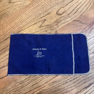 May include: A rectangular, navy blue pouch with white stitching around the edges. The text "STERLING BY POOLE" is printed in white, along with a small illustration of a ship and the text "A TRADITION IN SILVER SINCE 1893."