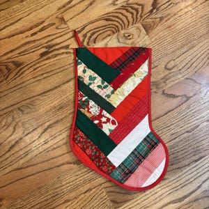 Può includere: Una calza natalizia trapuntata con un design patchwork diagonale. Le strisce di tessuto includono rosso e verde a tinta unita, tartan rosso, oro metallizzato, bianco e varie stampe a tema natalizio con agrifoglio, omini di pan di zenzero.