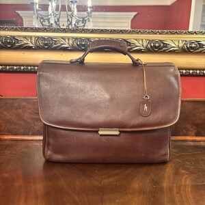 May include: A brown leather briefcase with a top handle and a metal clasp. The briefcase has a flap closure and a small leather tag with a letter "h" on it. The briefcase is sitting on a wooden surface.