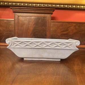 Puede incluir: Una jardinera rectangular de cerámica azul claro con un patrón de celosía en relieve en los laterales y detalles decorativos de volutas en cada extremo. El recipiente tiene un acabado liso, una base plana y una amplia abertura superior.