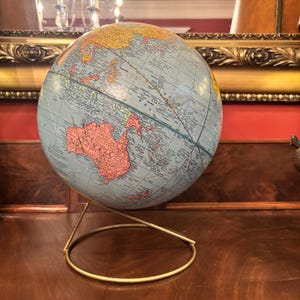 May include: A spherical world globe featuring a light blue ocean background with continents depicted in shades of yellow, orange, and pink. The globe is mounted on a thin, circular gold-toned metal stand that supports the sphere at an angle.