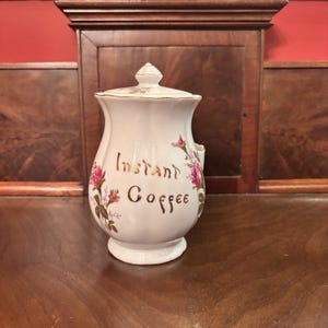 Op de afbeelding: Witte keramische instantkoffiepot met deksel, gouden rand en bloemmotieven. De woorden "Instant Coffee" zijn in gouden letters geschreven. De pot heeft een handvat en is versierd met roze rozen en groene bladeren.
