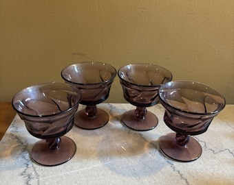 Op de afbeelding: Vier vintage amethistkleurige dessertglazen. Elk glas heeft een ronde kom met een textuurontwerp en staat op een korte steel en een cirkelvormige basis. De glazen zijn gerangschikt op een lichtgekleurd oppervlak.