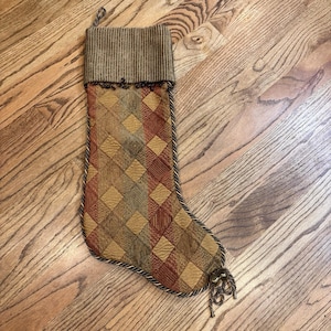 May include: A Christmas stocking with a diamond pattern in shades of brown, red, and gold. The top cuff is a ribbed brown fabric. The stocking is trimmed with a twisted rope-like border and has a loop for hanging.