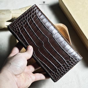 May include: A dark brown leather wallet with a crocodile skin texture. The wallet is rectangular and held in a hand. The surface has a textured appearance. The brand name "MESSIEURS" is visible at the top. The wallet is a rich, dark brown colour.