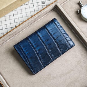 Puede incluir: Cartera de cuero azul marino con textura de piel de cocodrilo. La cartera rectangular está sobre una superficie gris claro. La cartera es de un tono azul oscuro con acabado brillante. La marca "MESSTEUR" es visible.