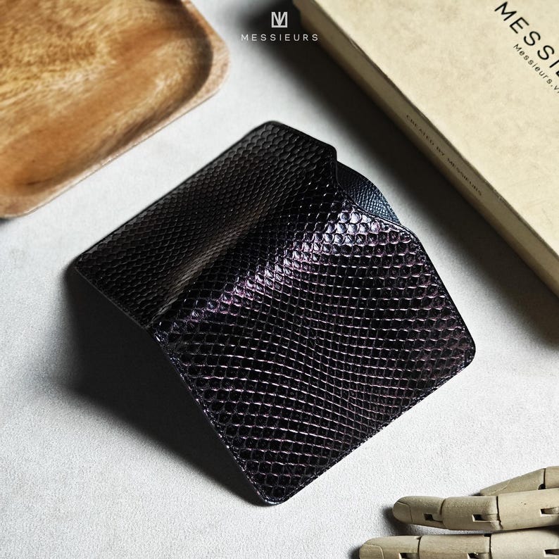 May include: A black, textured leather wallet with a reptile-like pattern. The wallet is open, revealing a glimpse of the interior. The Messieurs logo is visible on the top left, with a box in the background.
