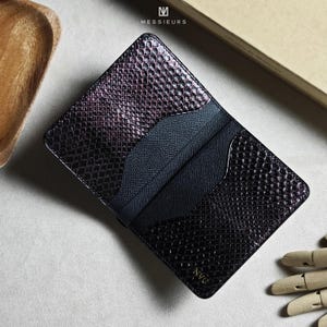 May include: A black and purple snakeskin-textured cardholder with multiple card slots. The cardholder has the word "Messieurs" printed on the top and the word "Nvo" printed on the bottom. The cardholder is open, revealing the interior.