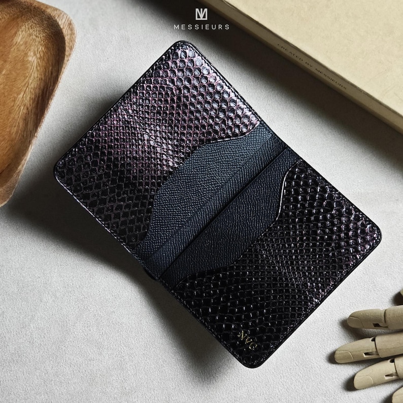 May include: A black and purple snakeskin-textured cardholder with multiple card slots. The cardholder has the word "Messieurs" printed on the top and the word "Nvo" printed on the bottom. The cardholder is open, revealing the interior.