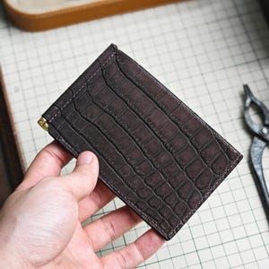 以下が含まれることがあります： クロコダイル柄の型押しが施されたダークブラウンの長方形の財布。財布の側面には、紙幣をしっかりと保持するための小さなゴールドトーンの金属製マネークリップが付いています。縁は同色の糸で丁寧に縫製されています。