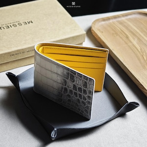 Puede incluir: Una cartera plegable con un exterior con textura de cocodrilo en tonos grises y blancos, con un interior amarillo brillante. La cartera se exhibe en una bandeja de cuero gris, con una caja de cartón en el fondo.