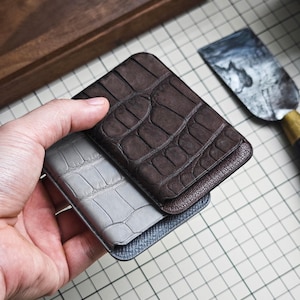 Puede incluir: Un tarjetero rectangular con una capa superior de cuero marrón oscuro con textura de cocodrilo y una capa inferior de cuero gris claro con textura de cocodrilo. La base del tarjetero tiene un acabado gris oscuro con textura cruzada.