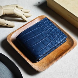 May include: A deep blue bifold wallet featuring a textured crocodile skin pattern. The wallet is square-shaped with visible stitching along the edges and displays the brand name MESSIEURS with a logo mark printed above it on the surface.
