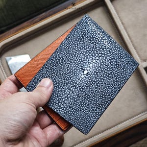 Puede incluir: Una cartera plegable con un exterior de cuero de raya texturizado gris oscuro y un interior de cuero naranja en contraste. La cartera se sostiene abierta, revelando el interior y el patrón único de la piel de raya.