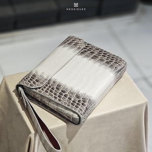May include: A rectangular clutch bag with a crocodile skin pattern in white and grey. The bag features a wrist strap and a flap closure. The brand name "MESSIERS" is visible in the top left corner.