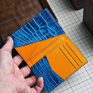 May include: A bi-fold wallet featuring a vibrant blue crocodile-embossed leather exterior and a bright orange textured leather interior. The interior includes multiple vertical card slots and a diagonal pocket, all secured with precise grey stitching.