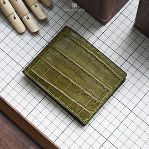 May include: A bi-fold wallet in an olive green colour featuring a textured crocodile skin pattern. The wallet has visible stitching along the edges and displays the brand name MESSIEURS in white text at the top centre of the product surface.