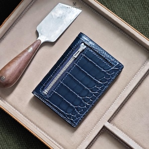 Op de afbeelding: Een marineblauwe leren portemonnee met een krokodillenleertextuur en een zilveren ritssluiting. De portemonnee is rechthoekig en heeft een strak design. Een metalen gereedschap met een houten handvat staat op de achtergrond. De merknaam "MESSIEURS" is zichtbaar.