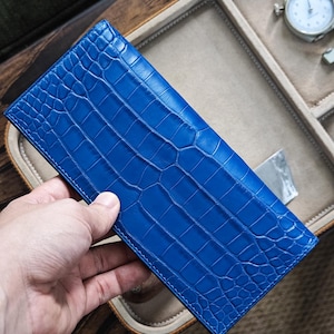 May include: A bright blue wallet with a crocodile skin pattern. The rectangular wallet is held by a hand. The brand name "MESSIERS" is visible. The background includes a watch and a storage tray. The wallet's dimensions are approximately 20 cm in length.