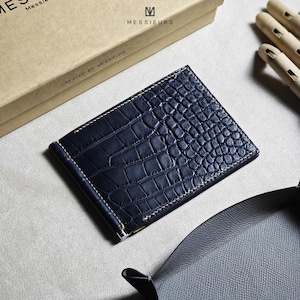 May include: A navy blue money clip wallet with a crocodile skin texture. The wallet has a metal clip and is stitched with yellow thread. The brand name "MESSIEURS" is visible on the packaging.